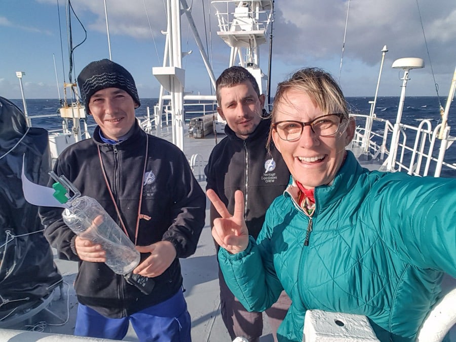 Three people stand on the deck of a research vessel under a partly cloudy sky, smiling and posing for a photo. One person holds a clear cylindrical sample container with green caps and a data tag, while another flashes a peace sign, suggesting a moment of celebration or camaraderie during a marine research mission.