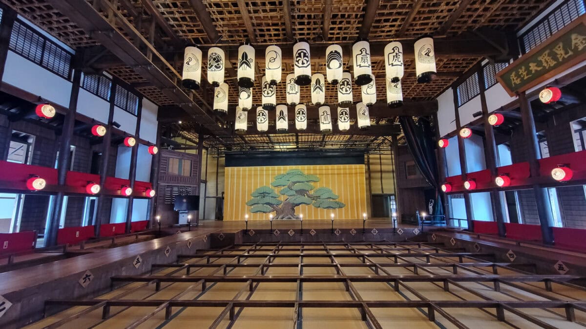 A traditional Japanese kabuki theater  stage on Shikoku Island with a vibrant yellow and green backdrop featuring a pine tree illustration. The auditorium is adorned with numerous hanging lanterns bearing Japanese symbols and characters, and red lanterns line the seating area. The space exudes a historic and cultural ambiance, perfect for an immersive performance experience.